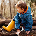 A young boy in Classic Wellies Yellow climbs a log in a forest. He's wearing a blue patterned jacket, and the sunlight dapples the scene.